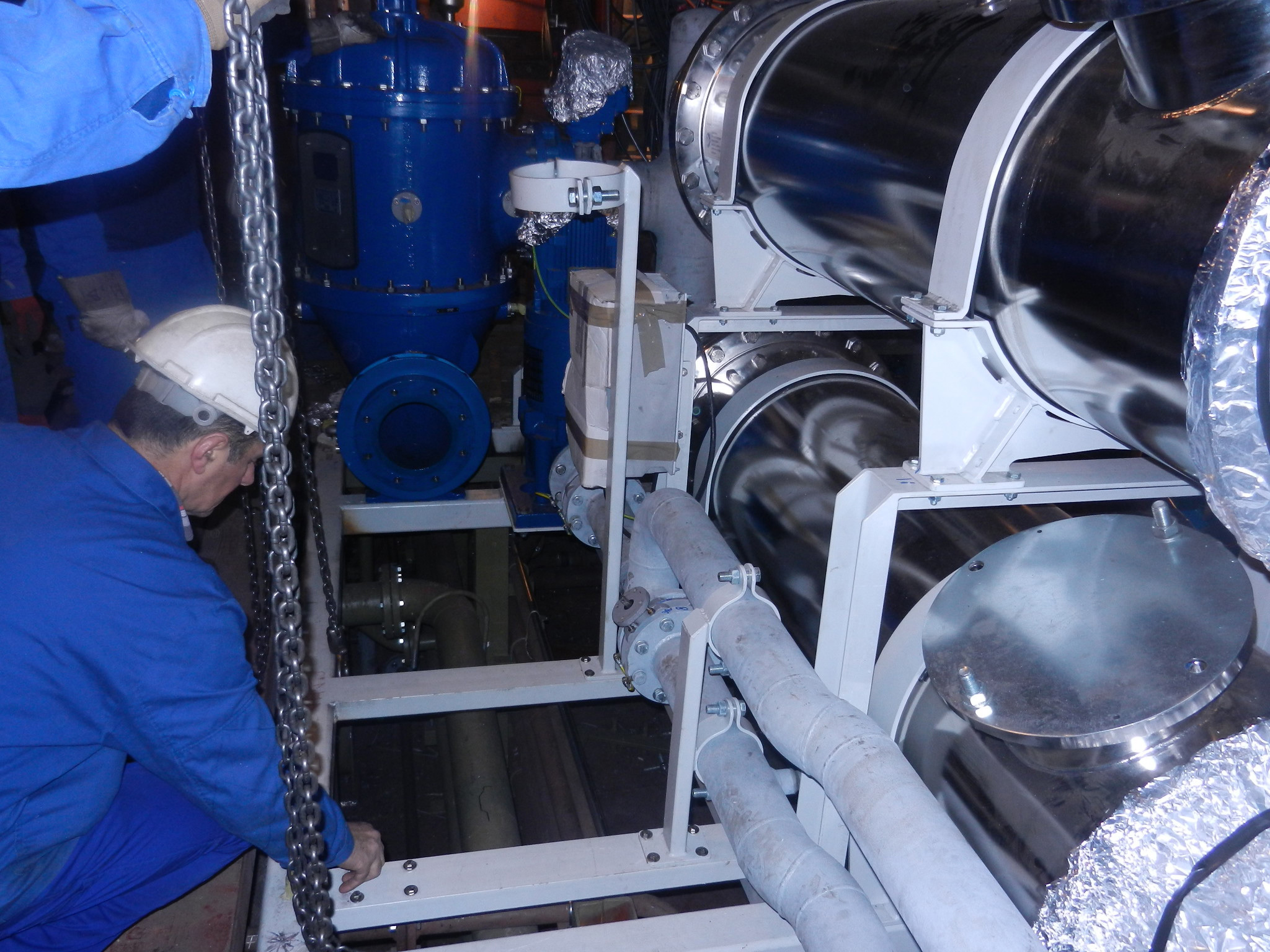 Worker in a hard hat adjusts industrial equipment with chains and pipes.