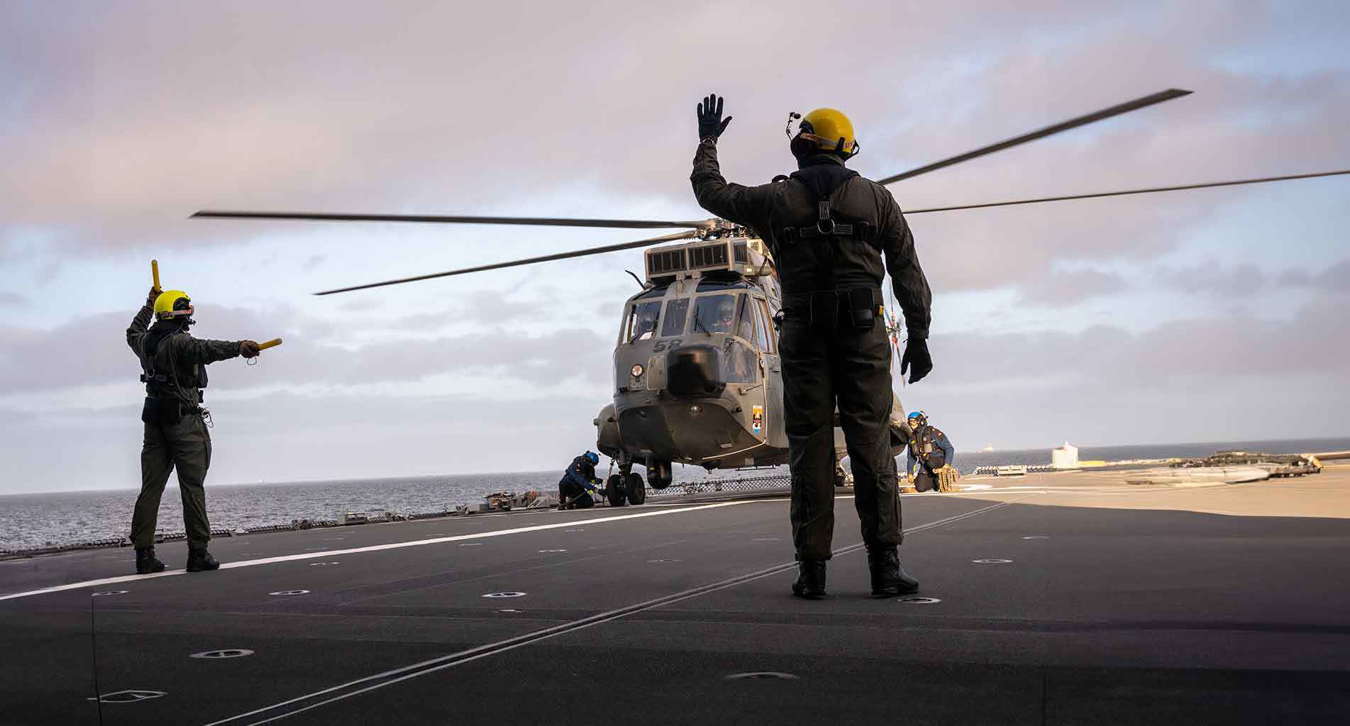 Helikopter landt op het dek van een vliegdekschip met bemanning die de piloot begeleidt.
