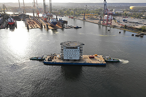 Large barge with a structure on a river near an industrial shipyard.