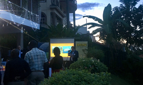 Outdoor presentation at dusk with attendees, projector screen, and illuminated decorations.