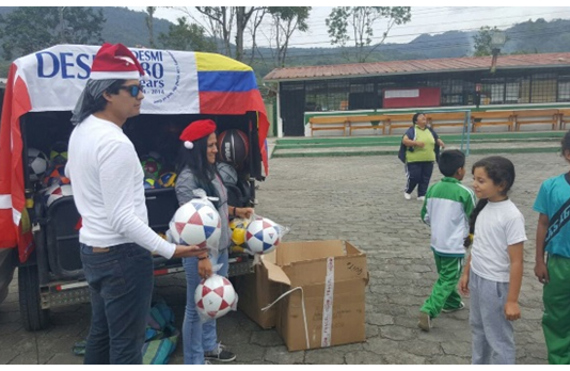 People distributing soccer balls to children in a schoolyard, with DESMI banner visible.