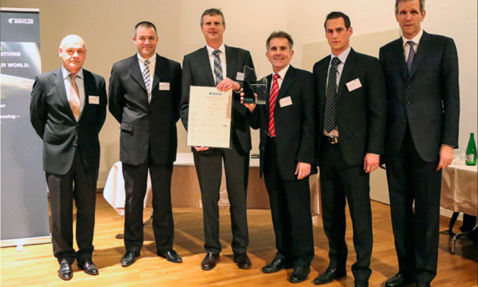 Six men in suits posing with an award and certificate in a formal setting.
