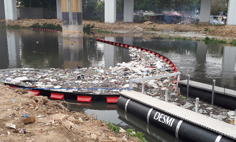 DESMI boom collecting debris and waste in a polluted river under a bridge.