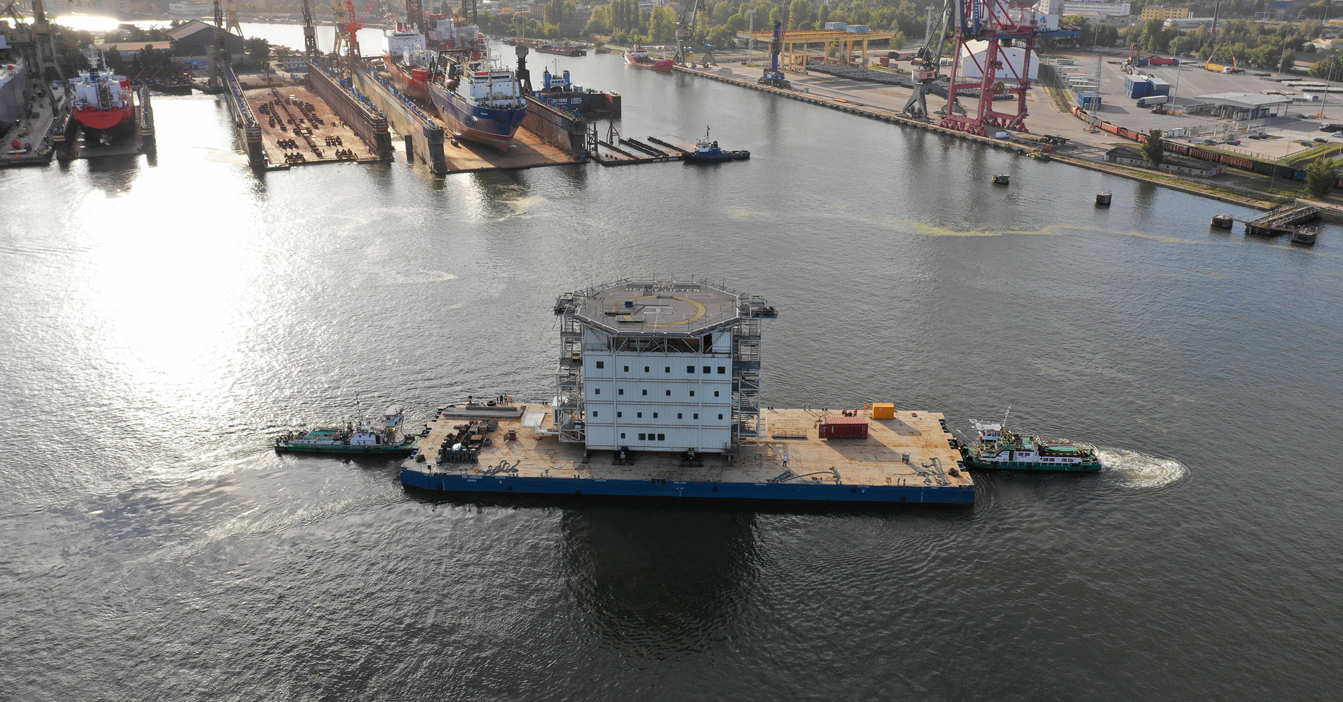 Large barge with helipad transported by tugboats in a busy shipyard.