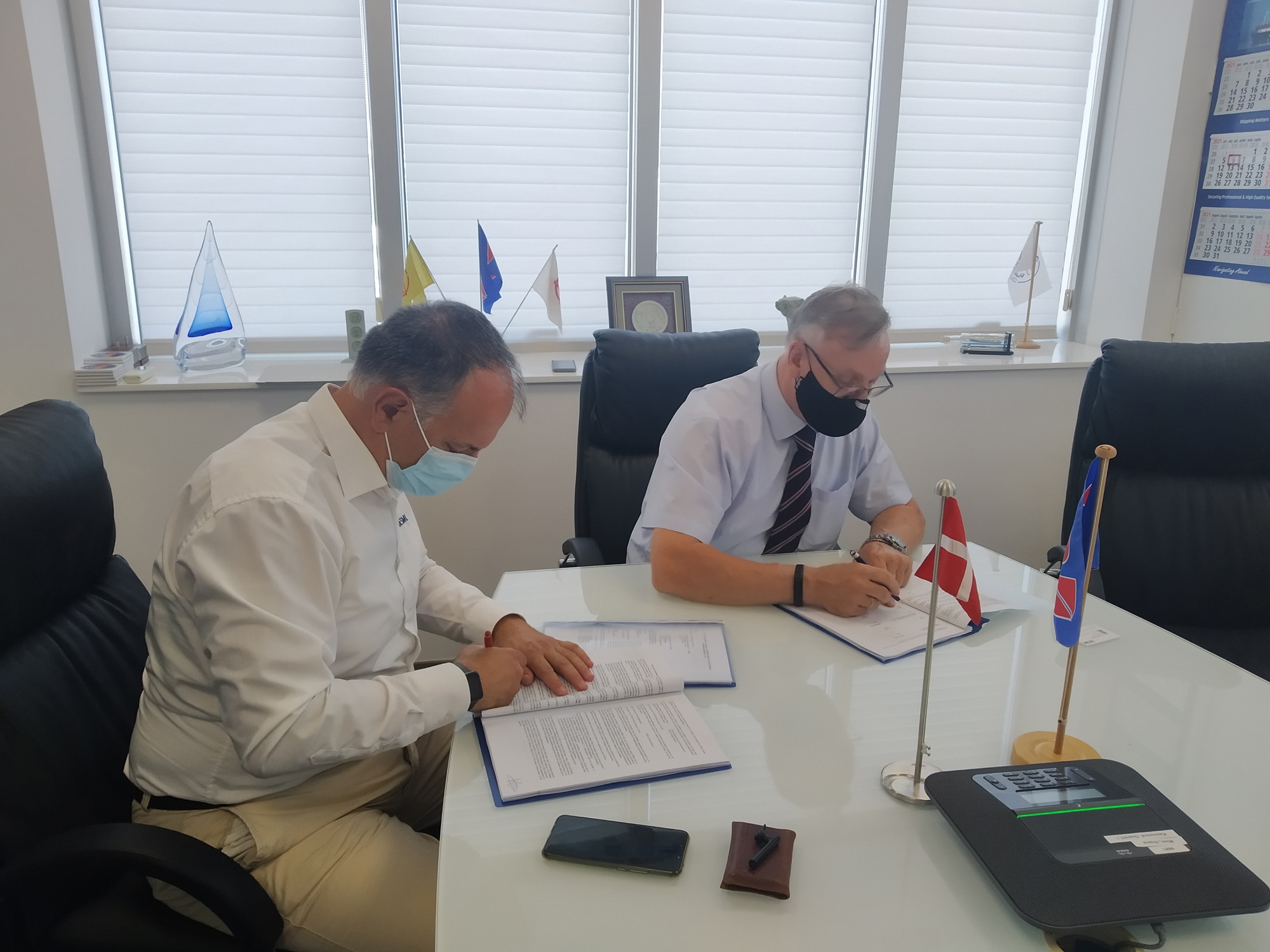Two men wearing masks signing documents at a conference table with flags.