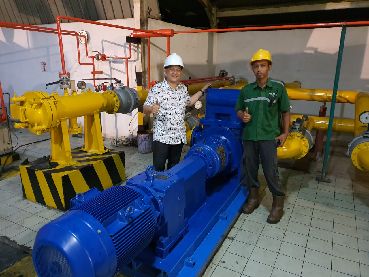 Two workers give thumbs up beside industrial pump in a mechanical room.
