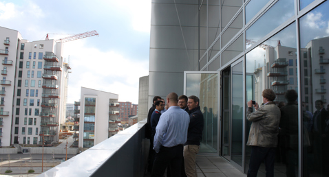 People conversing on a balcony with modern buildings and cranes in the background.