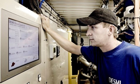 Technician monitors DESMI Ocean Guard A/S control panel in industrial setting.