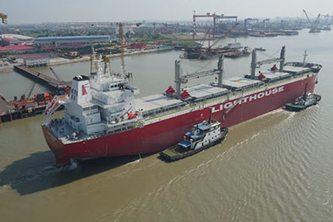 Red cargo ship "Lighthouse" in a busy industrial port with cranes.