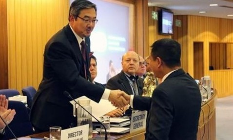Officials shaking hands at a formal meeting, with "Secretary General" nameplate visible.