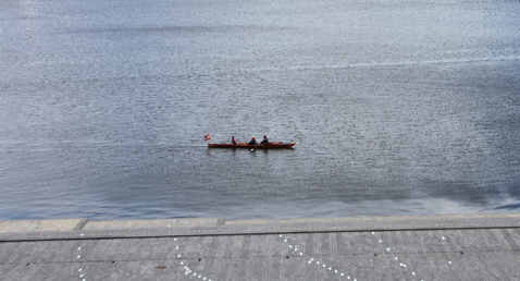 Three people rowing a boat with a Danish flag on a calm river.