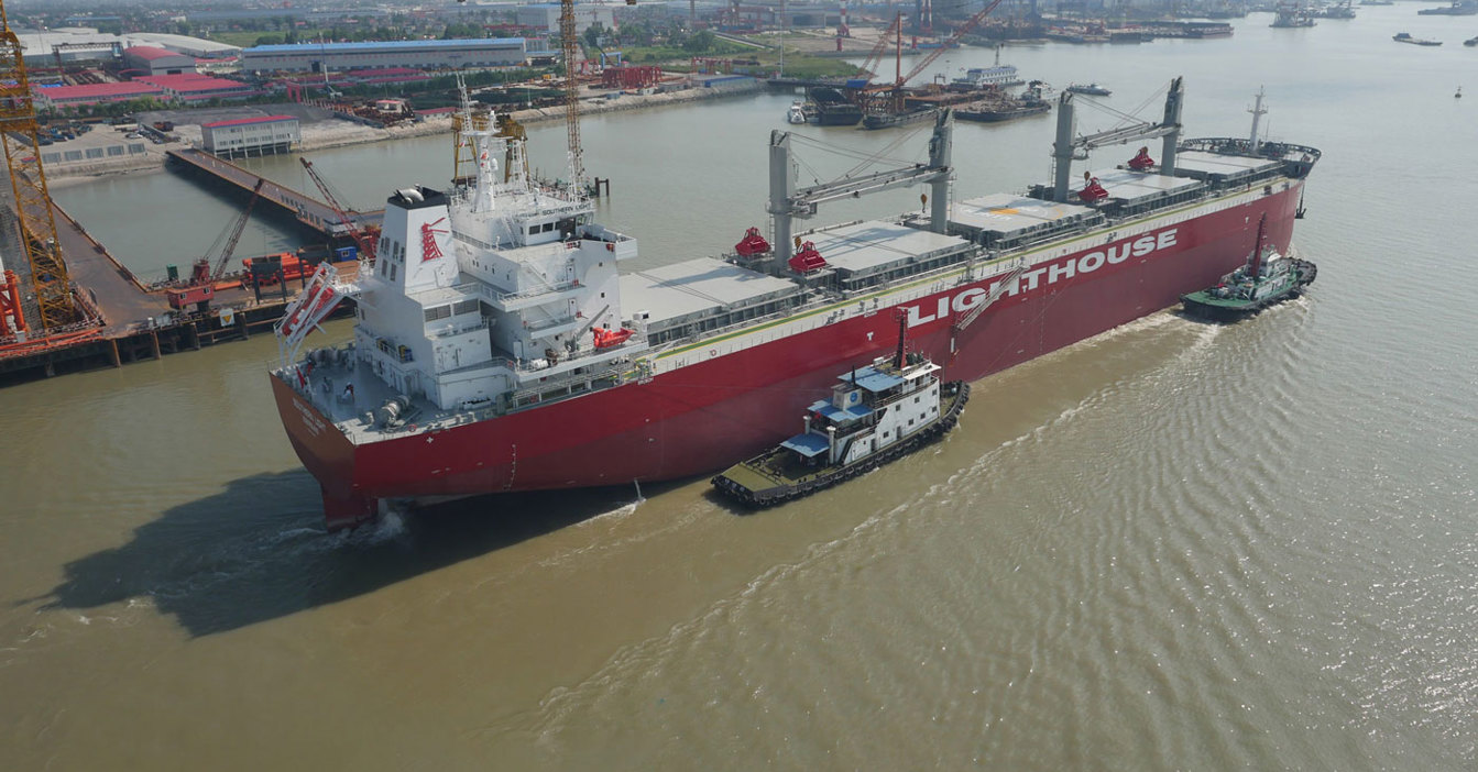 Large red cargo ship "Lighthouse" navigated by tugboats in a busy shipyard.