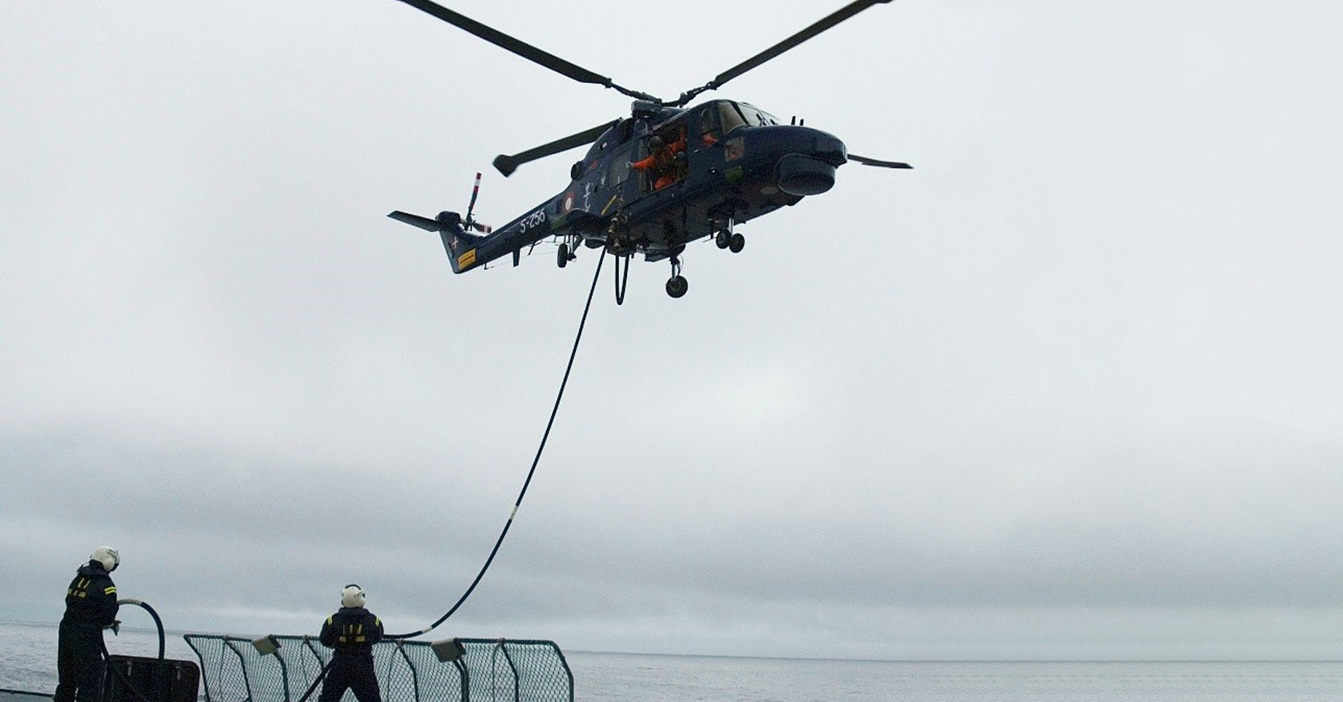 Helicopter refueling mid-air from ship, with crew managing fuel hose.