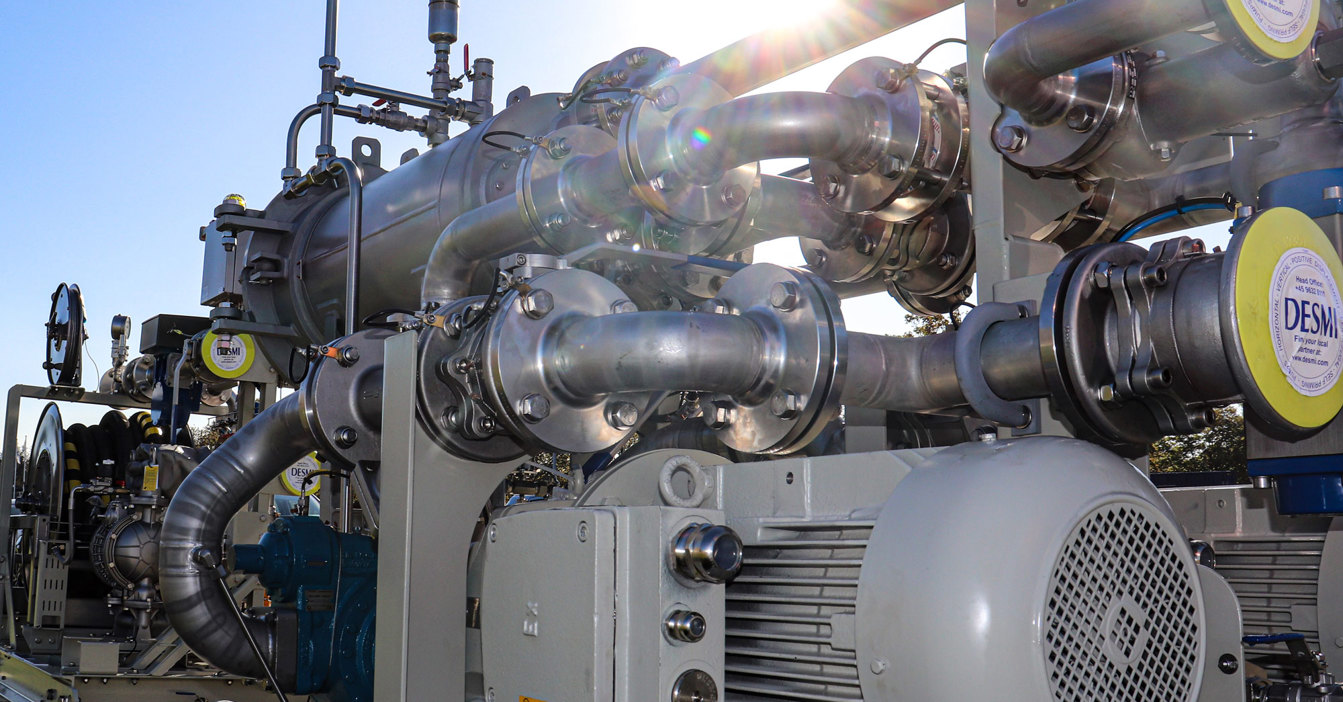 DESMI industrial pump system with pipes and valves, highlighted by sunlight.