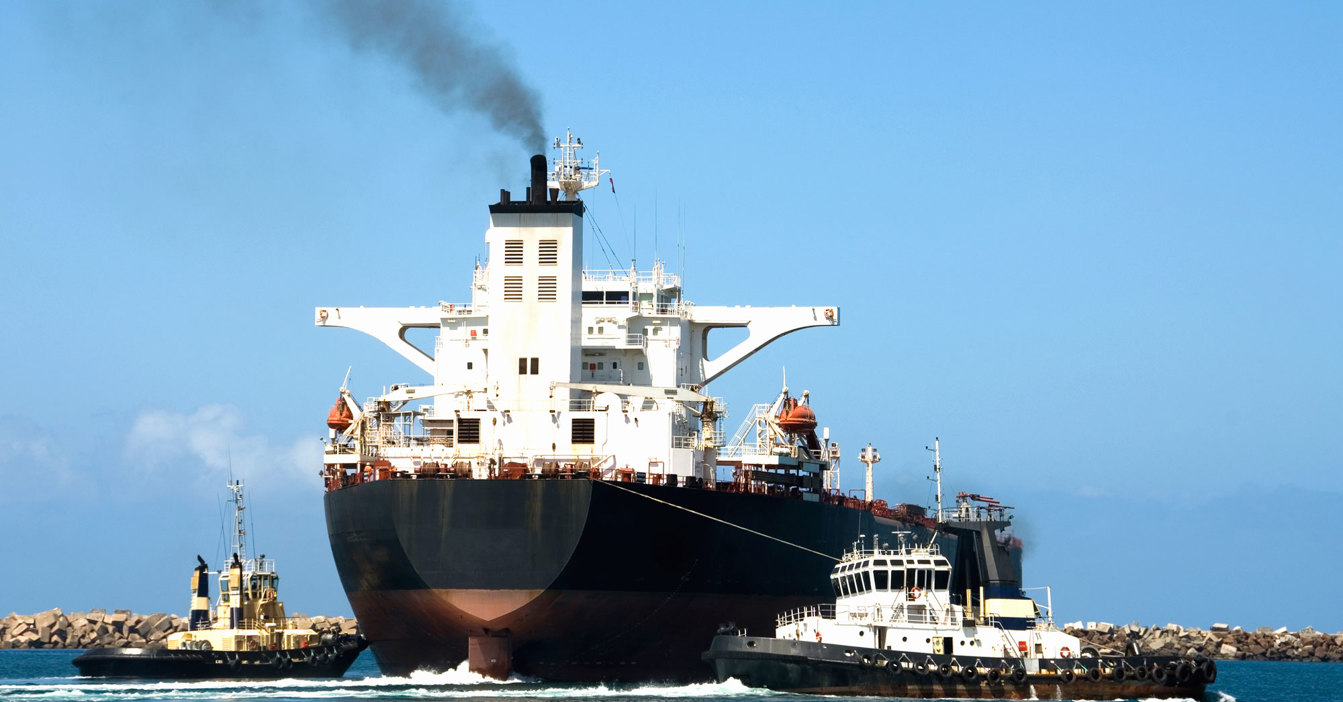 Container ship escorted by two tugboats, emitting smoke, in a harbor setting.