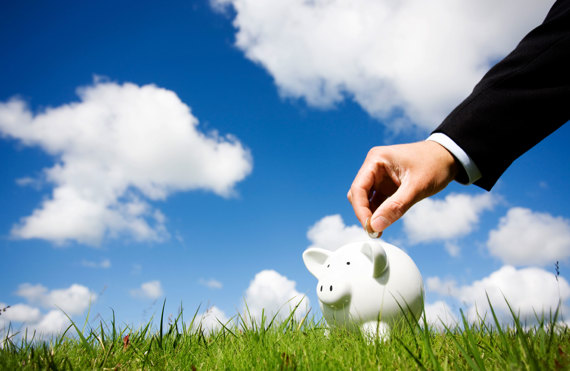 Hand placing coin into piggy bank on grass under blue sky with clouds.