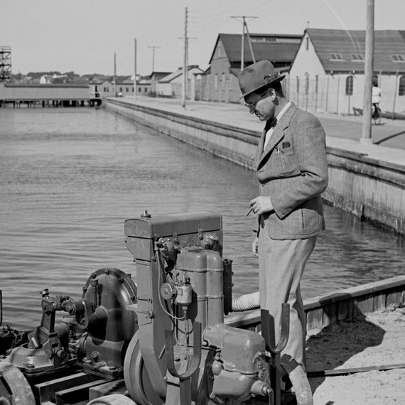 Man in suit inspecting industrial equipment by a waterfront.