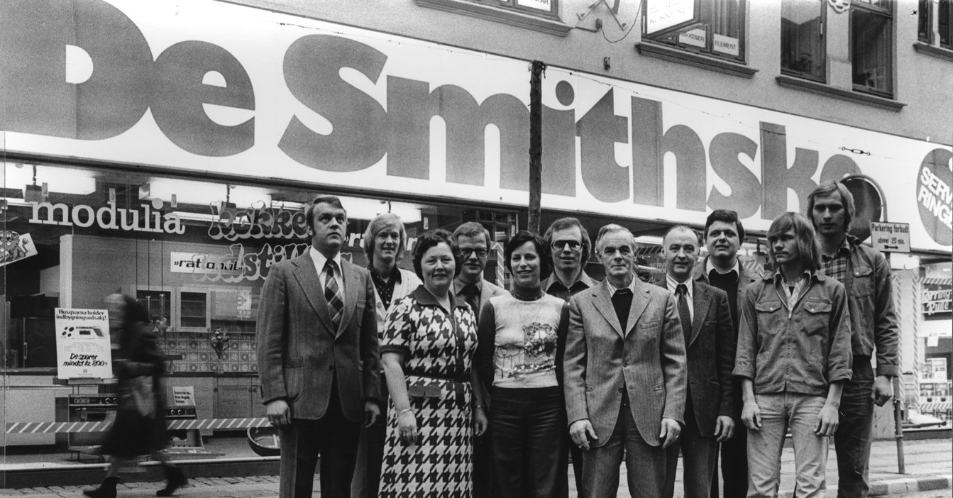Group of people posing in front of De Smithske store sign, black and white photo.