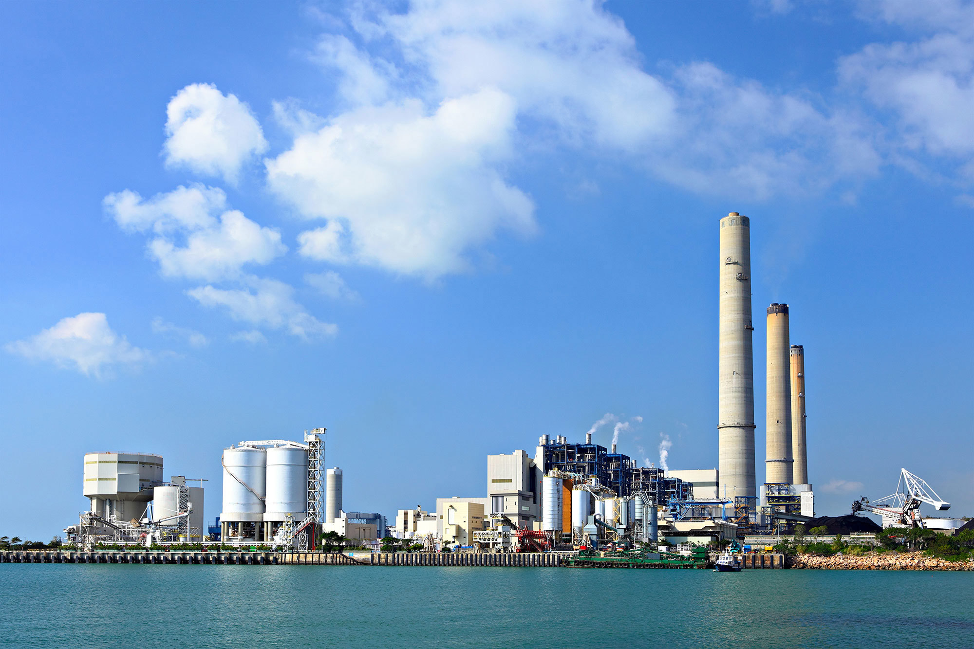 Industriellt kraftverk med höga skorstenar nära vatten under en klarblå himmel.