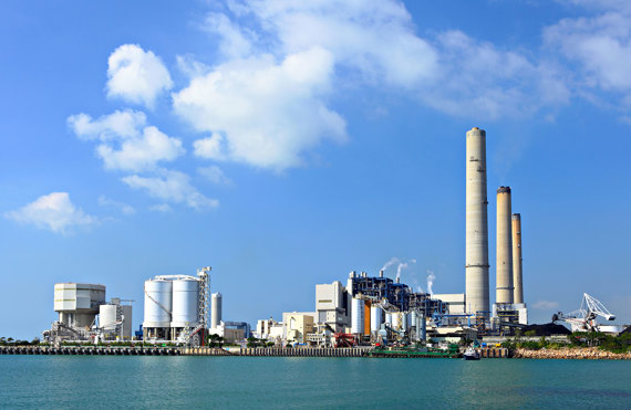 Industrial power plant with tall smokestacks near water under a clear blue sky.