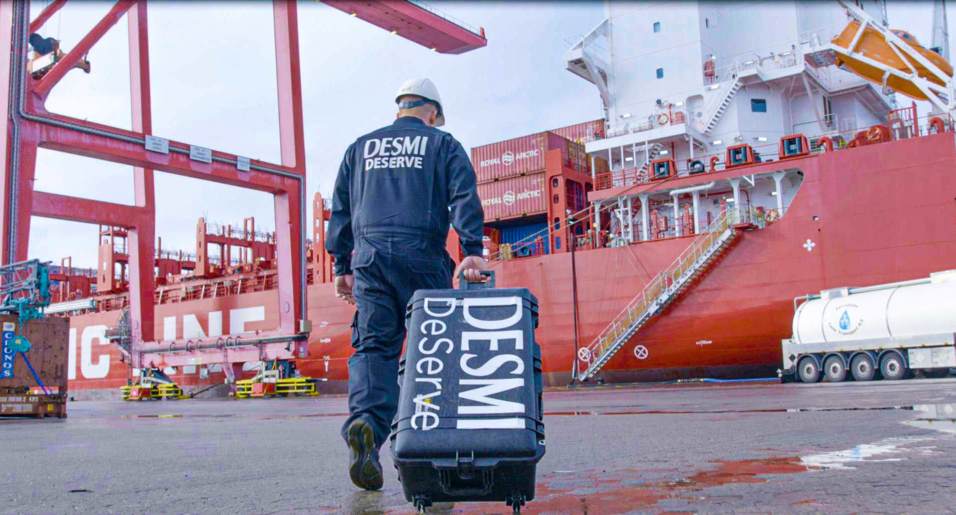 Worker in DESMI attire walks with equipment near a large red cargo ship.
