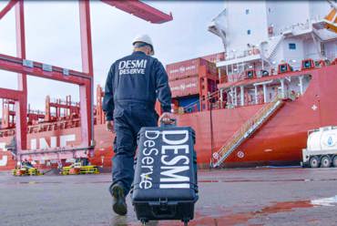 Worker in DESMI attire walks with equipment near a large red cargo ship.