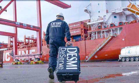 Worker in DESMI attire walks with equipment near a large red cargo ship.