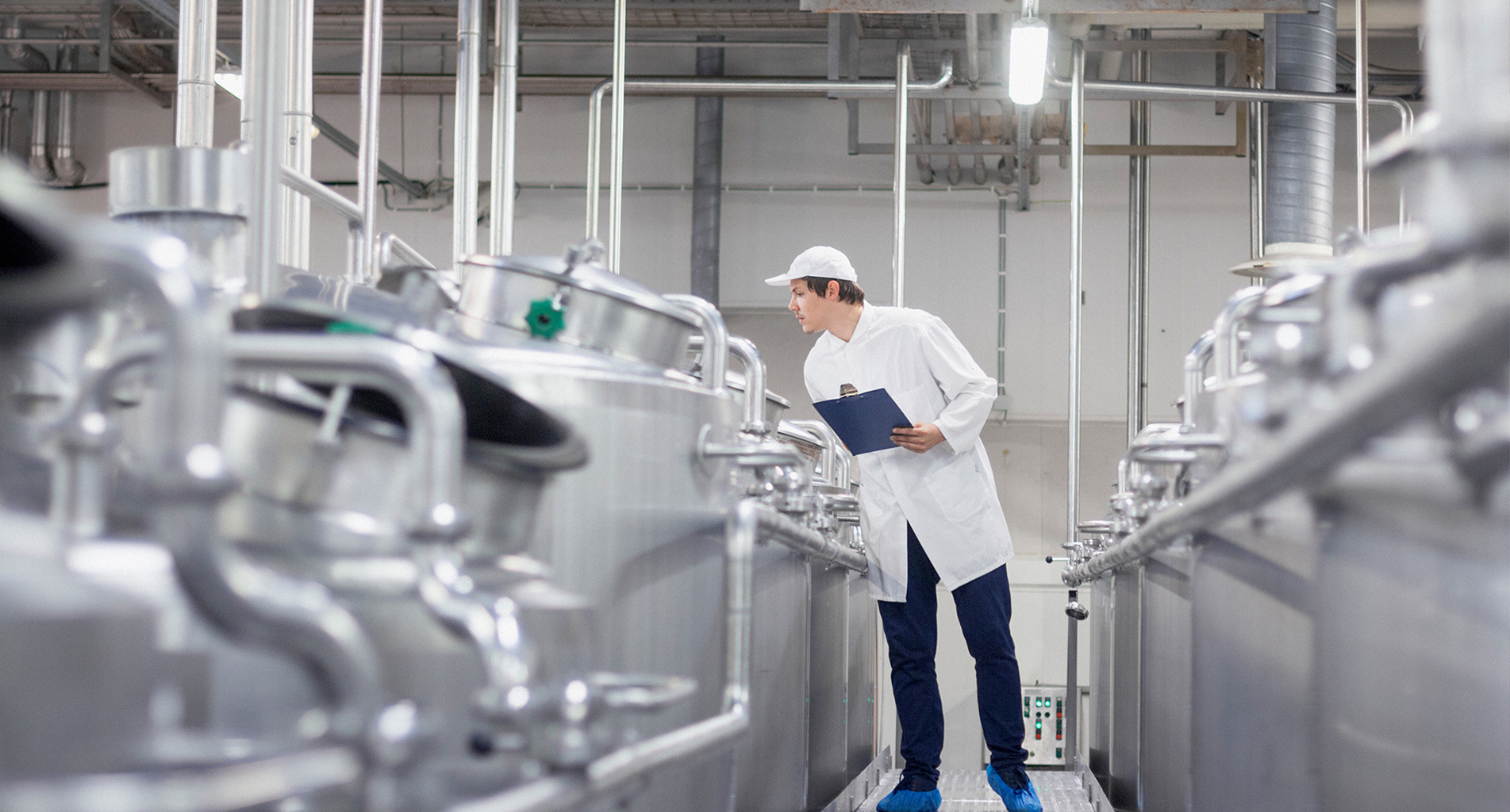 Trabajador inspeccionando tanques de acero inoxidable en una instalación industrial, sosteniendo un portapapeles.