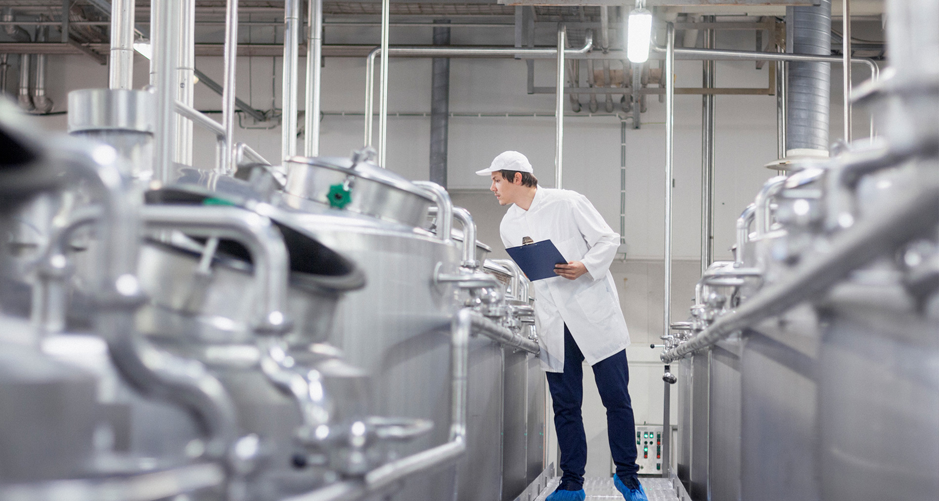 Travailleur inspectant des cuves en acier inoxydable dans une installation industrielle, tenant un clipboard.