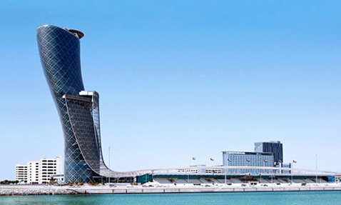 Modern, leaning skyscraper against a clear blue sky, reflected in calm water.