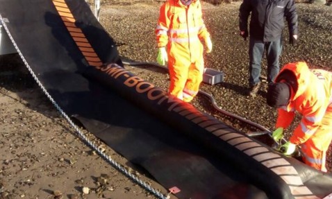 Workers deploying DESMI oil containment boom on rocky shoreline.