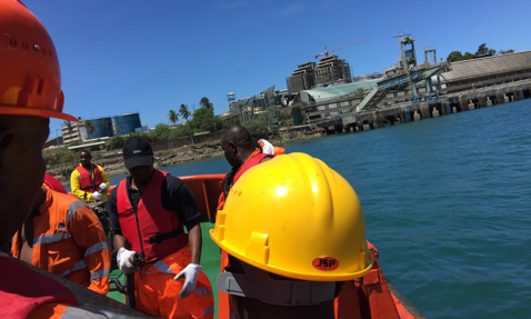Workers in safety gear on a boat near an industrial facility on the water.