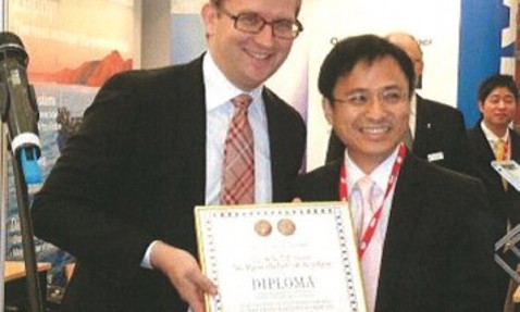 Two men smiling, holding a diploma at an event or exhibition.