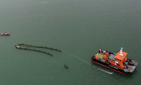 Aerial view of a vessel deploying an oil containment boom at sea.