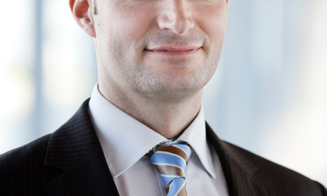 Smiling man in a suit and striped tie, with a blurred background.