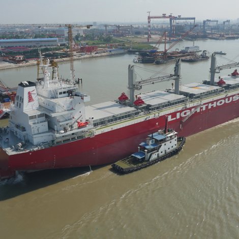 Large red cargo ship "Lighthouse" assisted by tugboats in a busy shipyard.