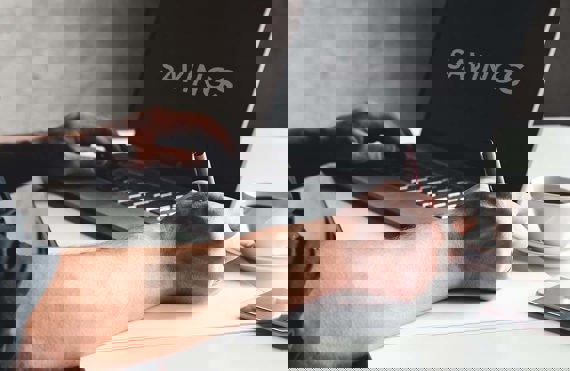 Person typing on laptop with "SAVINGS" on screen, holding a pen, coffee nearby.