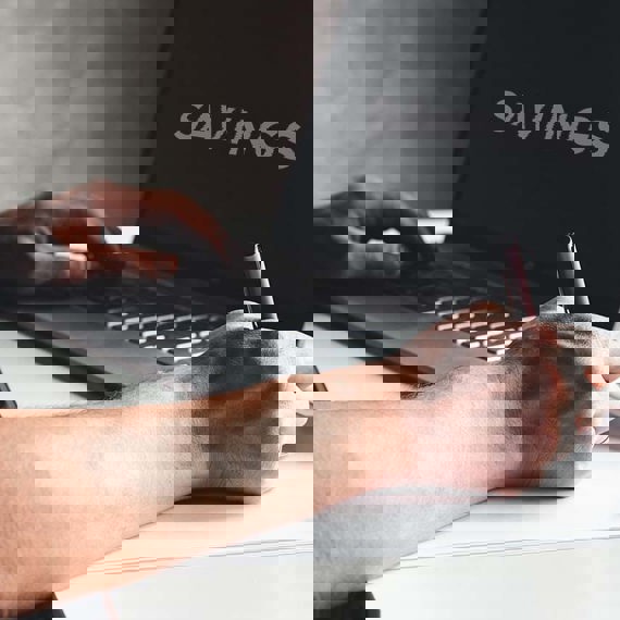 Persona escribiendo en una computadora portátil con "SAVINGS" en la pantalla, sosteniendo un bolígrafo, café cerca.
