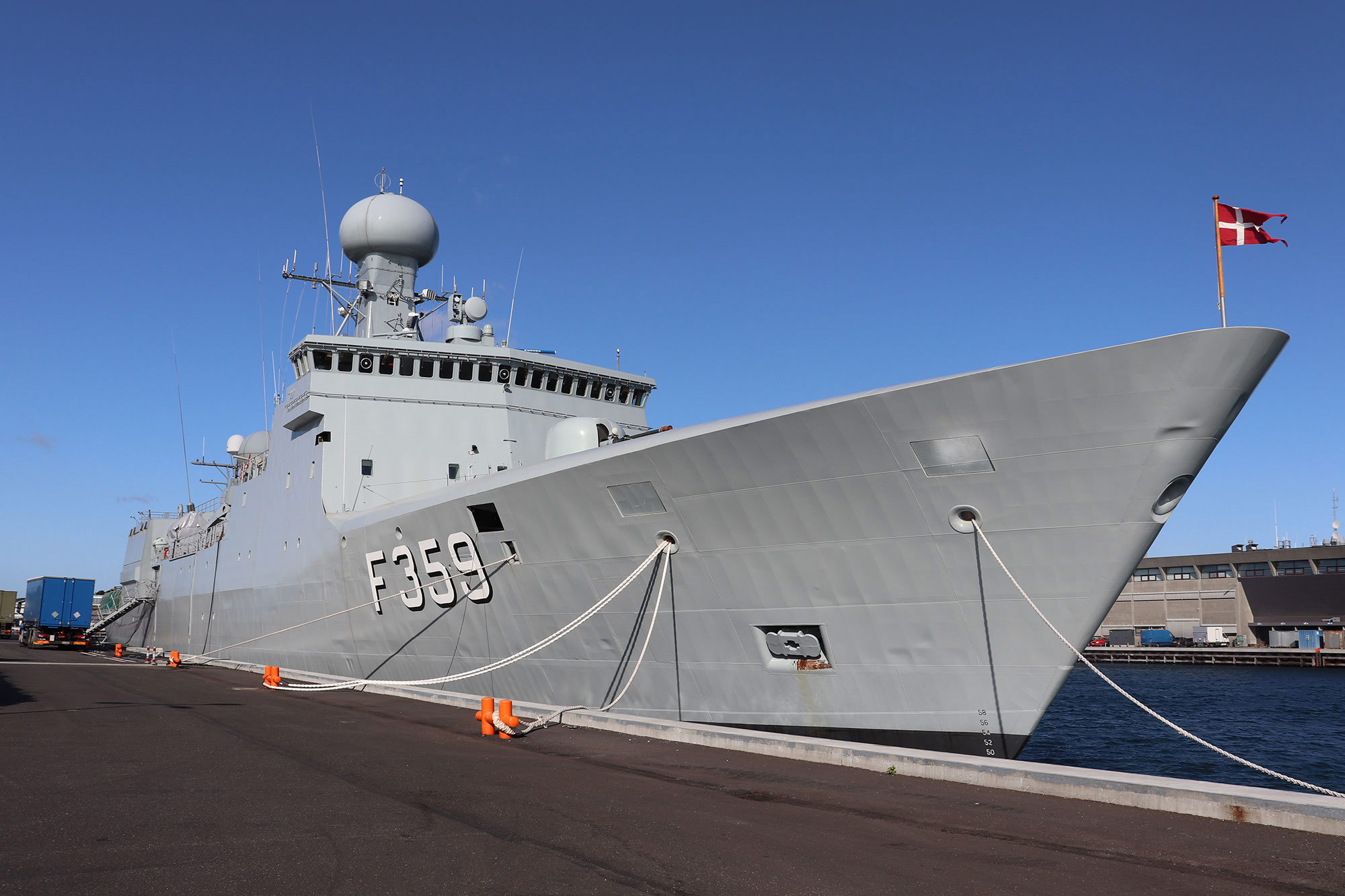 A naval ship docked at a pier, displaying the Danish flag.