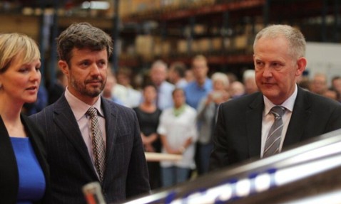 Three people in formal attire at an indoor event with a crowd.