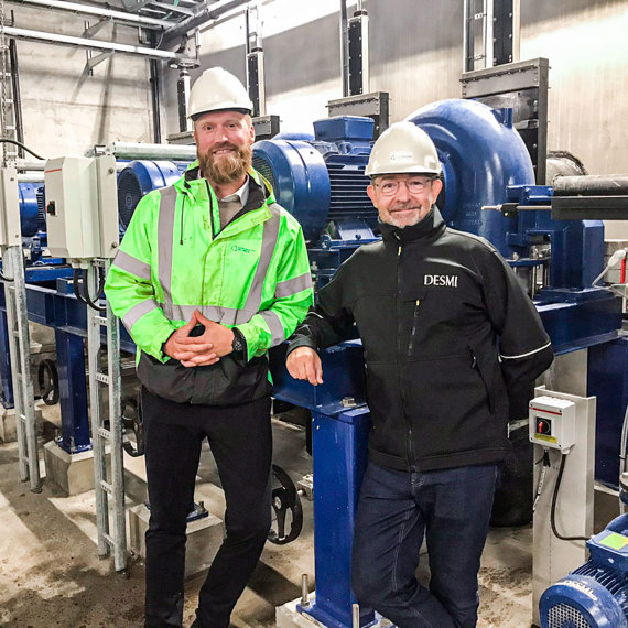 Two men in hard hats stand by DESMI industrial pumps in a facility.