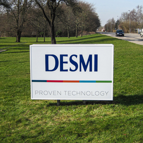 DESMI sign on grass with trees and road in background.