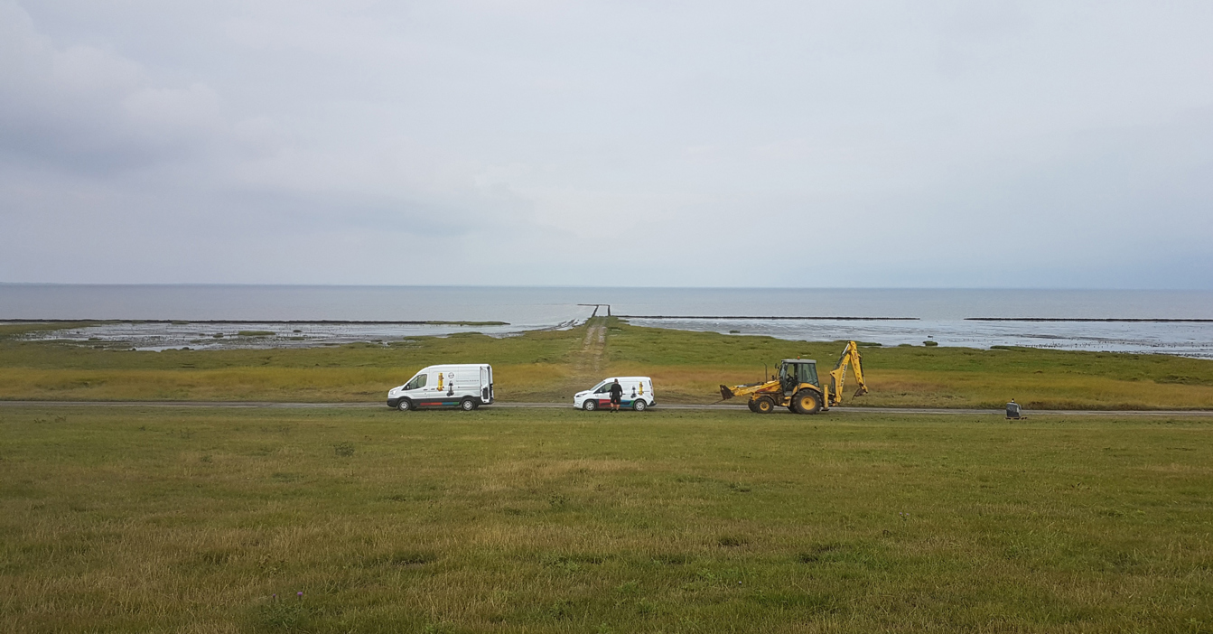 Vehicles and a backhoe on a coastal road under a cloudy sky.
