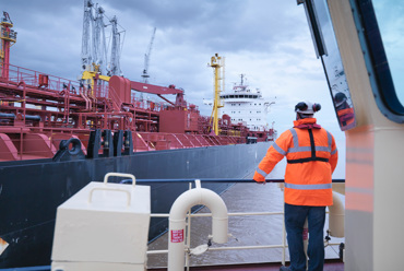 Worker in orange jacket observes large red industrial ship at port.