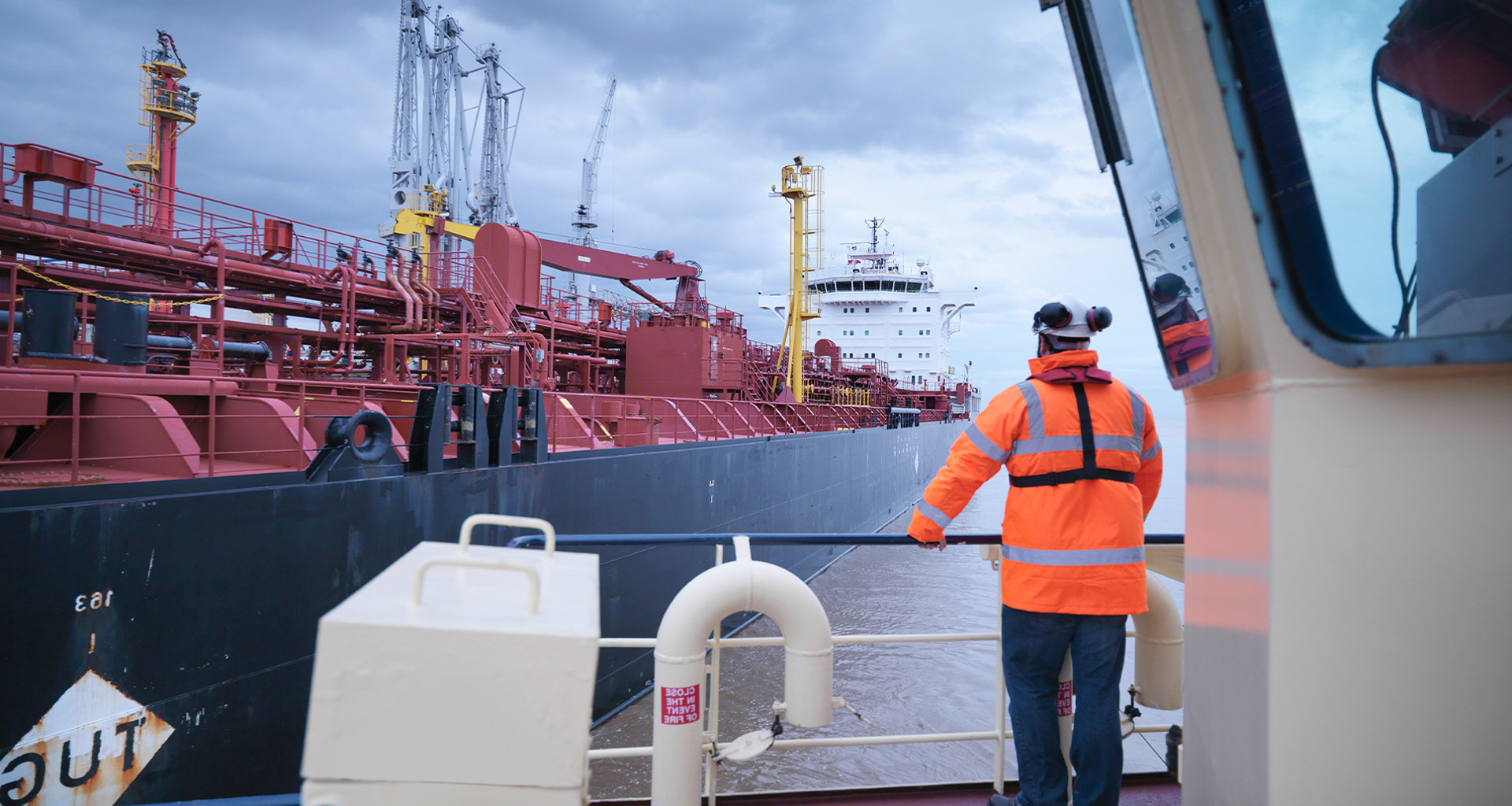 Travailleur en veste orange observe un grand navire industriel rouge au port.