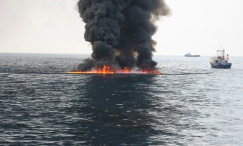 Thick black smoke and flames on ocean surface, with boats nearby.