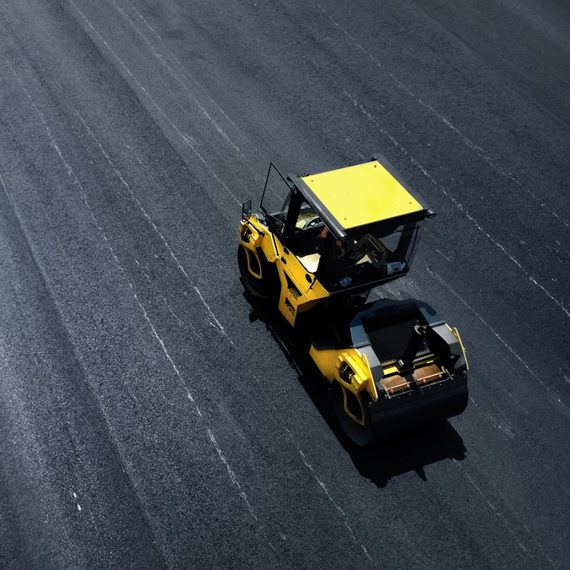 Yellow road roller on freshly paved asphalt, next to a white barrier.