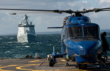 Blue helicopter on a ship's deck with a naval ship approaching.