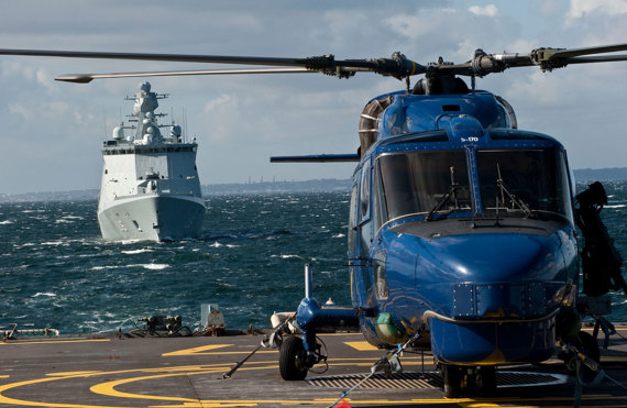 Blue helicopter on a ship's deck with a naval ship approaching.
