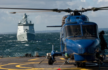 Blå helikopter på ett fartygsdäck med ett marinfartyg som närmar sig.